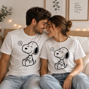 Puede incluir: Dos camisetas blancas con un dise&ntilde;o de dibujos animados de Snoopy en negro, cada una con un bocadillo en forma de coraz&oacute;n. Las camisetas las lleva una pareja, ambos con vaqueros azul claro. La pareja sonr&iacute;e y se toca la frente.