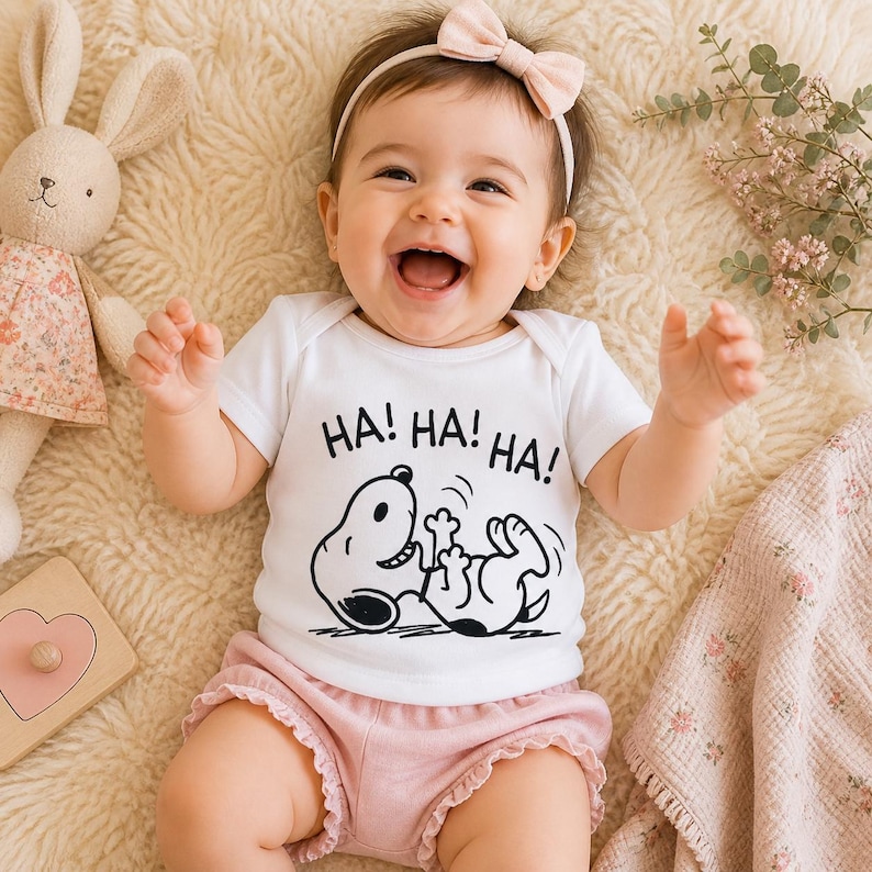Puede incluir: Un beb&eacute; con un body blanco con el texto "HA! HA! HA!" y un dibujo animado en blanco y negro de un perro riendo. El beb&eacute; tambi&eacute;n lleva un pantal&oacute;n corto rosa con volantes y una diadema rosa con lazo. Un conejito de peluche est&aacute; en el fondo.