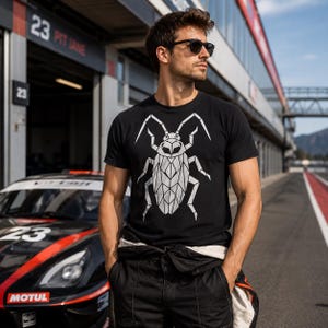 Op de afbeelding: Een zwart T-shirt met korte mouwen en een grote witte geometrische insectenafbeelding op de borst. Het shirt wordt gedragen met een zwarte broek en een donkere zonnebril, staand voor een zwart-rode raceauto bij een pitstraat op een circuit.