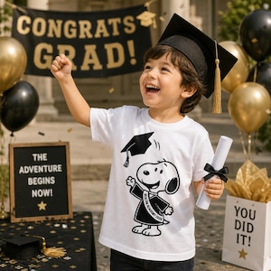 Puede incluir: Un ni&ntilde;o con birrete y camiseta blanca con un dise&ntilde;o de Snoopy y el texto "BEACON ACADEMY 2026". El fondo incluye globos, una pancarta que dice "CONGRATS GRAD!" y un letrero que dice "THE ADVENTURE BEGINS NOW!"