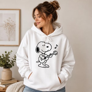 Puede incluir: Sudadera con capucha blanca con un gr&aacute;fico negro de Snoopy tocando la guitarra, con una nota musical encima. La sudadera tiene una capucha con cord&oacute;n y un bolsillo delantero. El dise&ntilde;o est&aacute; centrado en la parte delantera.