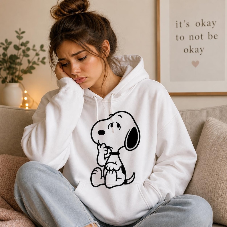Puede incluir: Sudadera con capucha blanca con un gr&aacute;fico en blanco y negro de Snoopy en una pose pensativa. La sudadera tiene una capucha con cord&oacute;n y un bolsillo de canguro. El texto en el arte de la pared dice "it's okay to not be okay".