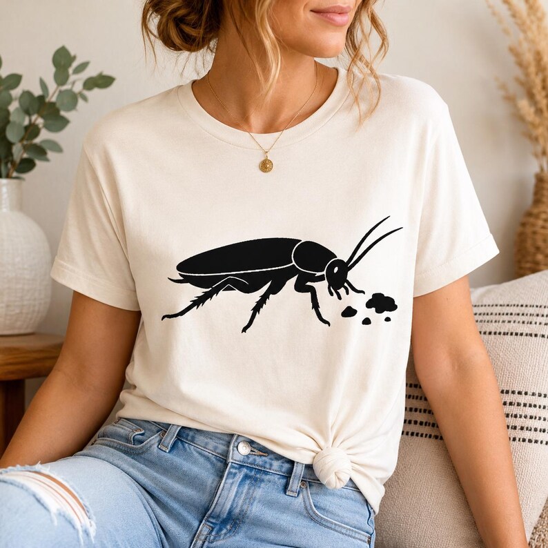 Op de afbeelding: Een cr&egrave;mekleurig T-shirt met korte mouwen en een zwarte grafische afbeelding van een kakkerlak op de voorkant. De illustratie van het insect toont het wezen in profiel met lange antennes en kleine stippen bij de mond.