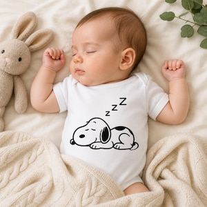 Puede incluir: Un beb&eacute; durmiendo con un body blanco con un dise&ntilde;o de Snoopy en blanco y negro. El beb&eacute; est&aacute; acostado sobre una manta color crema junto a un conejito de peluche. El body tiene la letra &laquo;Z&raquo; repetida tres veces encima del dise&ntilde;o de Snoopy.