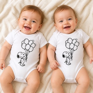 Puede incluir: Dos beb&eacute;s con bodies blancos con un dibujo negro de Snoopy sosteniendo globos. Los beb&eacute;s sonr&iacute;en y est&aacute;n acostados sobre una manta color crema. Los bodies son de manga corta.