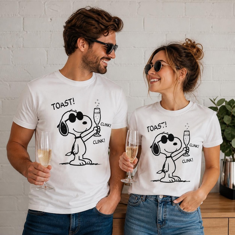 Puede incluir: Camisetas blancas con un dise&ntilde;o de dibujos animados de Snoopy en negro, con gafas de sol y sosteniendo una copa de champ&aacute;n. El texto "TOAST!" y "CLINK!" est&aacute;n impresos en las camisetas. Dos personas llevan las camisetas y sostienen copas de champ&aacute;n.