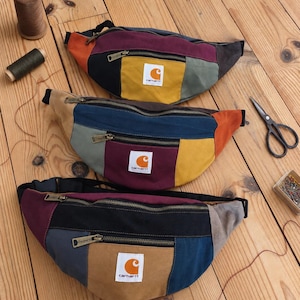 Pode incluir: Três pochetes color-block com um emblema do logótipo Carhartt no bolso frontal. Cada saco é construído a partir de vários painéis de tecido em tons de amarelo mostarda, bordeaux, azul marinho, preto, laranja e cinzento com fechos de metal.