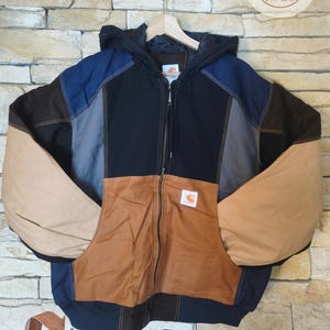 Puede incluir: Una chaqueta con capucha de bloques de color que presenta paneles de tela negra, tostada, azul marino y gris. La chaqueta tiene una cremallera frontal completa, una capucha y un parche de la marca Carhartt en blanco en el bolsillo delantero.
