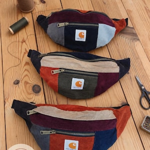 Pode incluir: Três pochetes coloridas feitas de tecido de veludo cotelê, cada uma com um bolso frontal com zíper e um emblema da marca Carhartt. As bolsas estão dispostas verticalmente, exibindo vários painéis de tecido azul-marinho, bordô, laranja e cinza.