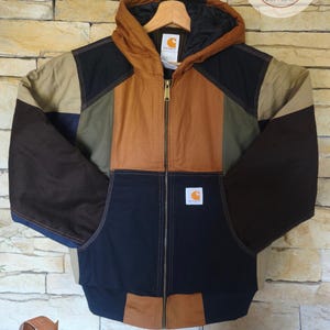 Può includere: Una giacca con cappuccio color-block caratterizzata da pannelli in tessuto marrone, beige, verde oliva scuro e blu navy. La giacca presenta una chiusura frontale con zip, un cappuccio e una toppa del marchio Carhartt sul davanti a sinistra.