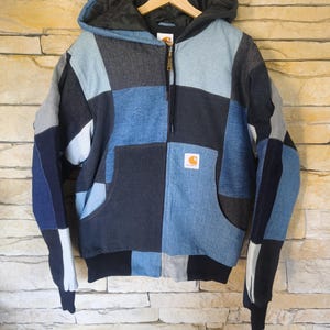 Peut inclure: Une veste à capuche zippée présentant un motif patchwork composé de divers panneaux de tissu bleu et gris rappelant le denim. La veste comprend une poche kangourou, une doublure de capuche noire, des poignets et un ourlet côtelés noirs.
