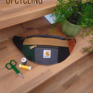 May include: A colour-blocked fanny pack featuring panels of black, dark green, tan, grey, and rust orange fabric with a Carhartt logo patch on the front pocket. The bag includes metal zippers, green sewing scissors, and two spools of thread.