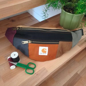 May include: A color-blocked fanny pack featuring panels of burgundy, charcoal gray, black, orange, and tan canvas. The bag has two zippered compartments with brass-toned hardware and a white Carhartt brand patch. Includes green scissors and thread.