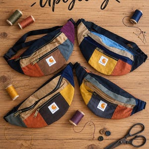 Puede incluir: Cuatro riñoneras de bloques de color hechas de tela reutilizada, cada una con un parche de la marca Carhartt, un bolsillo frontal con cremallera y una correa negra ajustable. La palabra Upcycling está escrita en letra negra arriba.