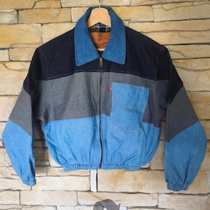 Könnte beinhalten: Eine farblich abgesetzte Jeansjacke mit dunkelblauem Kragen und Brustbereich, einem grauen Mittelteil und einem hellblauen unteren Bereich. Sie verfügt über einen Frontreißverschluss, eine Brusttasche mit rotem Etikett und elastische Ärmelbündchen.