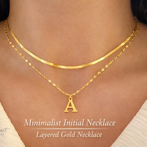 Puede incluir: Un collar dorado en capas con un diseño minimalista. La cadena superior es una banda plana y lisa, mientras que la cadena inferior presenta un diseño delicado y con cuentas. Un colgante dorado con la forma de la letra "A" cuelga de la cadena inferior. El texto "Minimalist Initial Necklace" es visible.