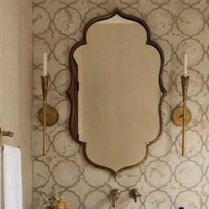 May include: A bathroom scene featuring a decorative mirror with an ornate gold frame, a stone sink, and gold sconces. The sink is set on a white marble countertop. A white towel hangs on a gold towel rack. The wall is decorated with a patterned wallpaper.