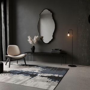 May include: A modern interior scene featuring a large, irregularly shaped mirror with a black frame. A black console table holds a vase with dried flowers and books. A patterned rug in shades of gray, blue, and black sits on the floor, with a chair and a floor lamp.