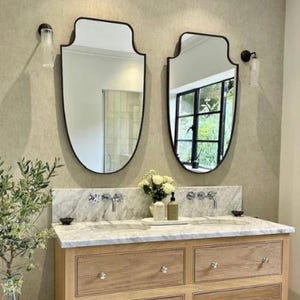May include: A double vanity featuring a light wood cabinet with four drawers and silver knobs, topped with a white marble countertop. Two shield-shaped mirrors with black frames are mounted above, flanked by two cylindrical wall sconces.