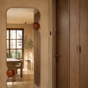 Pode incluir: Um espelho de parede alto e retangular com cantos arredondados e um perfil de borda irregular e ondulado. O espelho apresenta dois proeminentes detalhes decorativos circulares em castanho brilhante fixados na moldura do lado esquerdo, um no topo e outro na base.