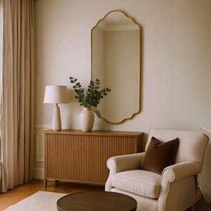 Peut inclure: Intérieur de salon avec un grand miroir doré orné. Un buffet en bois à rainures verticales, un fauteuil crème avec un coussin marron et une table basse ronde sont également présents. Une lampe et un vase avec de la verdure complètent le décor.