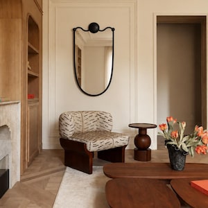 May include: A black framed mirror with a circular top detail, a low-profile armchair upholstered in a cream fabric with a black dash pattern, a dark wood pedestal side table, and a low-profile dark wood coffee table with orange tulips.