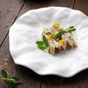 Może przedstawiać: Biały, organicznie ukształtowany talerz zawiera porcję pokrojonego kurczaka, udekorowanego małymi żółtymi i białymi kwiatami, zielonymi liśćmi i zieloną oliwką. Talerz stoi na ciemnej drewnianej powierzchni.