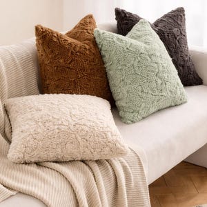 May include: A collection of fluffy throw pillows in various colors, including cream, brown, and green, arranged on a neutral-toned sofa. The pillows have a textured, shaggy appearance, adding a cozy element to the home decor.