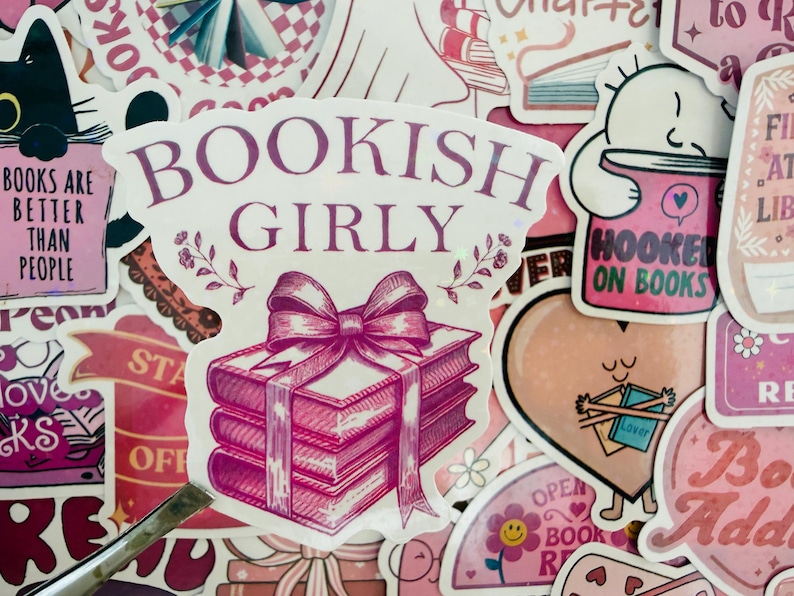 May include: A selection of pink and white stickers with book-related designs. One sticker says "Bookish Girly" and features a stack of books tied with a bow. Other stickers include phrases like "Books are better than people" and "Hooked on Books."