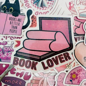 May include: A selection of pink and white stickers with book-related designs. One sticker shows a black cat reading a book with the text "BOOKS ARE BETTER THAN PEOPLE." Another says "BOOK LOVER" with a heart design. Other stickers have book and heart motifs.