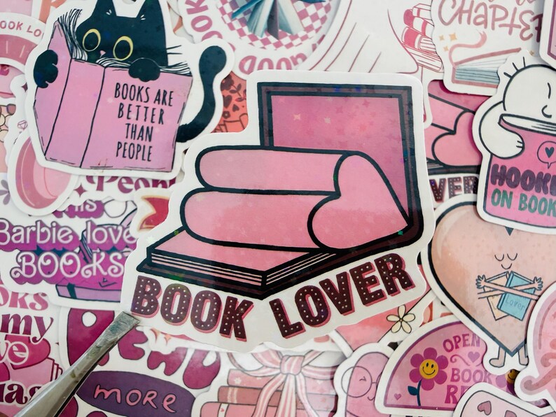 May include: A selection of pink and white stickers with book-related designs. One sticker shows a black cat reading a book with the text "BOOKS ARE BETTER THAN PEOPLE." Another says "BOOK LOVER" with a heart design. Other stickers have book and heart motifs.