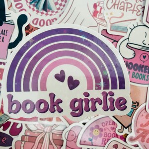 May include: A sticker with a rainbow design in shades of purple and pink, featuring two small hearts and the text "book girlie." The sticker is surrounded by other stickers with book-related themes and text.