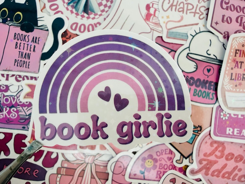 May include: A sticker with a rainbow design in shades of purple and pink, featuring two small hearts and the text "book girlie." The sticker is surrounded by other stickers with book-related themes and text.