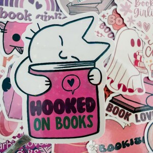 May include: A sticker depicting a white cat-like figure embracing a pink book, with the text "HOOKED ON BOOKS" in green and black. The image is surrounded by other stickers with book-related designs in pink and white.