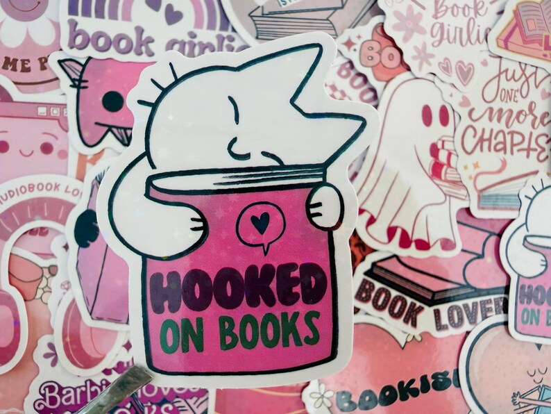 May include: A sticker depicting a white cat-like figure embracing a pink book, with the text "HOOKED ON BOOKS" in green and black. The image is surrounded by other stickers with book-related designs in pink and white.