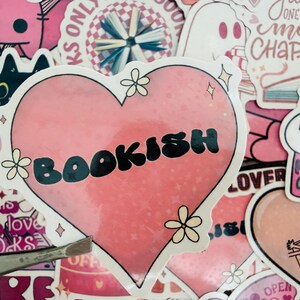 May include: A selection of pink stickers with a book theme. The main sticker is heart-shaped and says "BOOKISH" in black. Other stickers feature a ghost, a cat reading, and the text "HOOKED ON BOOKS".