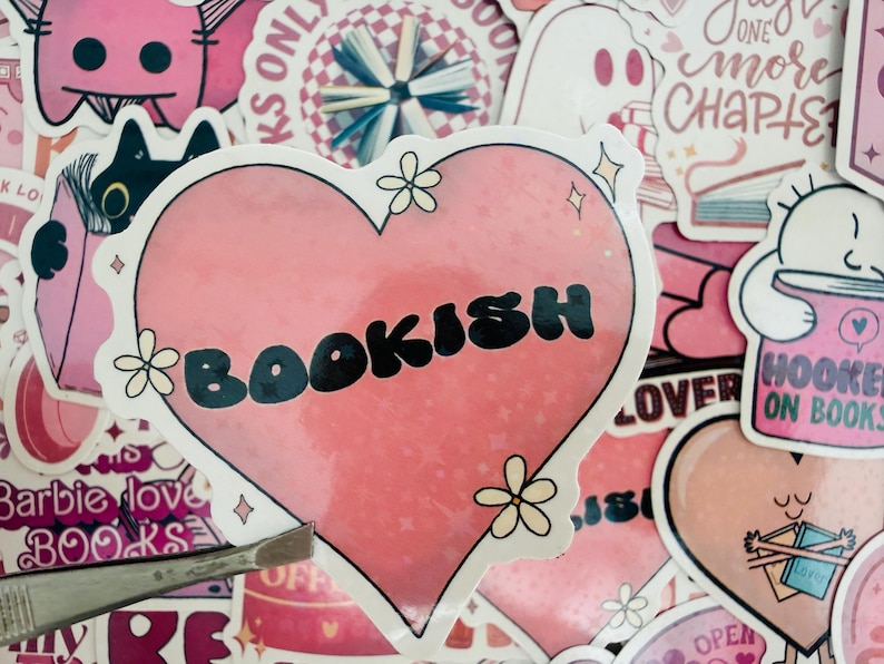 May include: A selection of pink stickers with a book theme. The main sticker is heart-shaped and says "BOOKISH" in black. Other stickers feature a ghost, a cat reading, and the text "HOOKED ON BOOKS".
