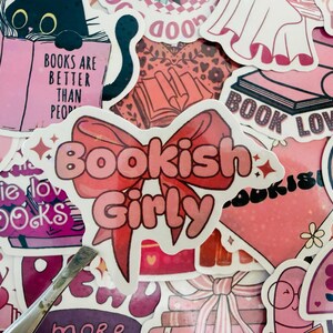 May include: A selection of pink stickers with book-related designs. One sticker says "Bookish Girly" with a bow. Other stickers show a black cat reading a book, the text "Books are better than people," and the words "Book Lover."