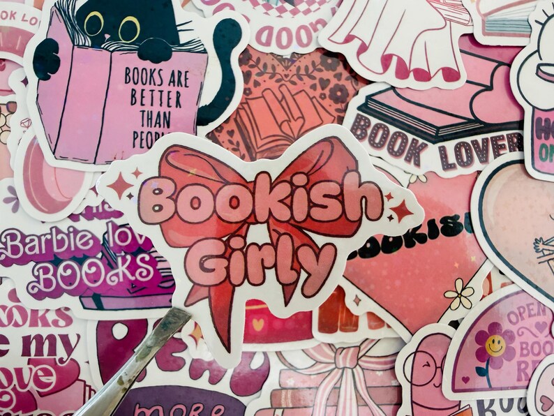 May include: A selection of pink stickers with book-related designs. One sticker says "Bookish Girly" with a bow. Other stickers show a black cat reading a book, the text "Books are better than people," and the words "Book Lover."