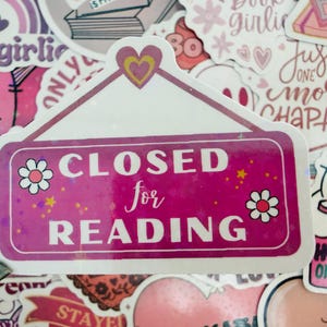 May include: A pink sticker with the text "CLOSED for READING" in white. The sticker features a white border, small flower and star decorations, and a heart at the top. Surrounded by other stickers.