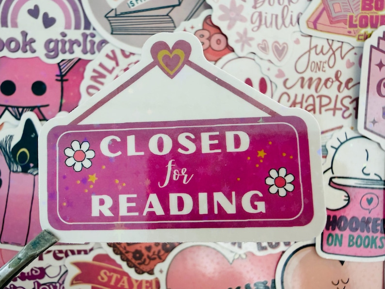 May include: A pink sticker with the text "CLOSED for READING" in white. The sticker features a white border, small flower and star decorations, and a heart at the top. Surrounded by other stickers.