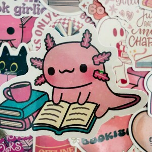 May include: A pink axolotl sticker reading a book, with a stack of books and a coffee cup. The sticker is surrounded by other pink and white stickers with book-related themes and text like "book girlie" and "Hooked on Books."