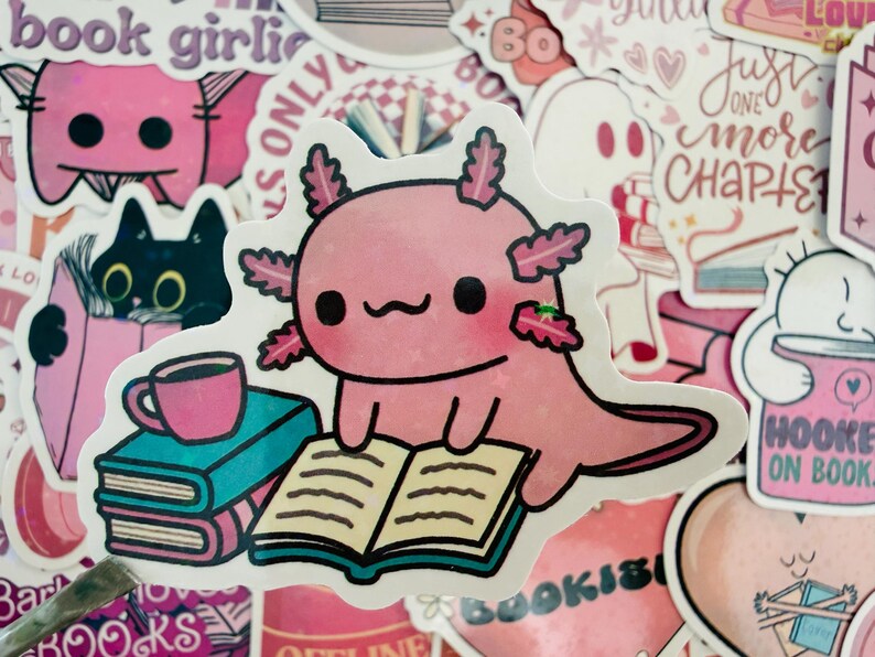 May include: A pink axolotl sticker reading a book, with a stack of books and a coffee cup. The sticker is surrounded by other pink and white stickers with book-related themes and text like "book girlie" and "Hooked on Books."