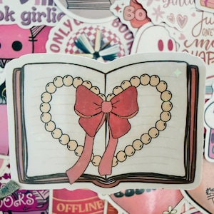 May include: A sticker depicting an open book adorned with a heart-shaped pearl necklace and a red bow. The sticker is surrounded by other pink stickers with text like "book girlie" and "Hooked on Books".