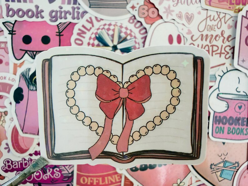 May include: A sticker depicting an open book adorned with a heart-shaped pearl necklace and a red bow. The sticker is surrounded by other pink stickers with text like "book girlie" and "Hooked on Books".