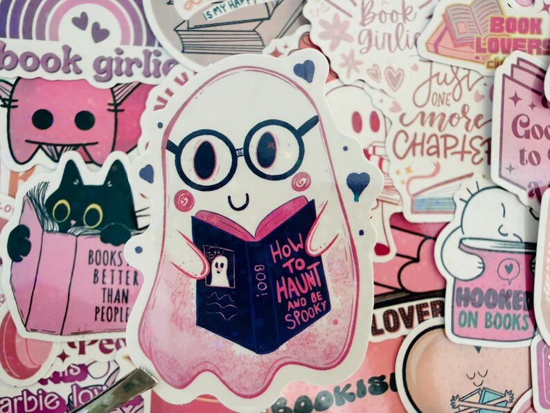 May include: A selection of pink and white stickers with a book theme. The main sticker shows a ghost wearing glasses and reading a book titled "How to Haunt and Be Spooky." Other stickers include a black cat reading, and text that says "Book Lovers."