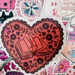 May include: A red heart-shaped sticker with a floral and book design. The sticker has a decorative border and features an open book illustration. Surrounding stickers have pink and white designs with text like "Hooked on Books" and "One More Chapter".