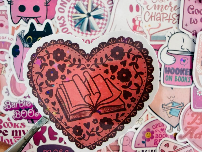 May include: A red heart-shaped sticker with a floral and book design. The sticker has a decorative border and features an open book illustration. Surrounding stickers have pink and white designs with text like "Hooked on Books" and "One More Chapter".