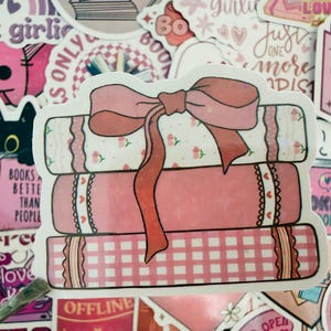 May include: A sticker depicting a stack of three pink books tied with a large pink bow. The books have decorative patterns, including floral and gingham designs, surrounded by other stickers with text and illustrations.
