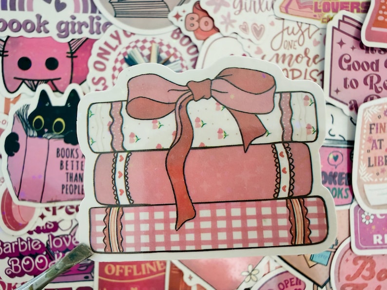 May include: A sticker depicting a stack of three pink books tied with a large pink bow. The books have decorative patterns, including floral and gingham designs, surrounded by other stickers with text and illustrations.